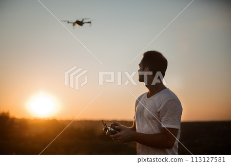 Silhouette of a man piloting drone in the air with a remote controller in his hands on sunset. Pilot takes aerial photos and videos with quadcopter from above 113127581