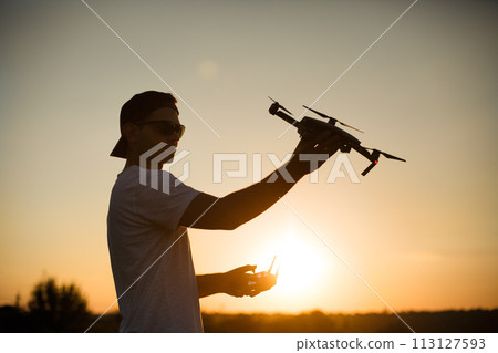 Silhouette of a man holding small compact drone and remote controller in his hands. Pilot launches quadcopter from his palm on sunset. Drone ready to take off. 113127593