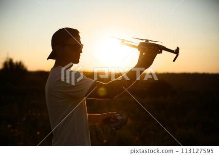 Silhouette of a man holding small compact drone and remote controller in his hands. Pilot launches quadcopter from his palm on sunset. Drone ready to take off. Silhouette of a man holding small compact drone and remote controller in his hands. Pilot launches quadcopter from his palm on sunset. Drone ready to take off. 113127595