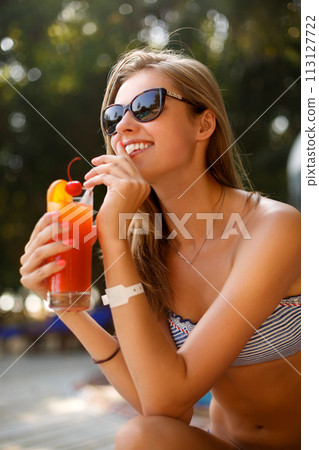 Portrait of young woman with cocktail glass chilling in the tropical sun near swimming pool on a deck chair with palm trees behind. Vacation concept Portrait of young woman with cocktail glass chilling in the tropical sun near swimming pool on a deck chair with palm trees behind. Vacation concept 113127722