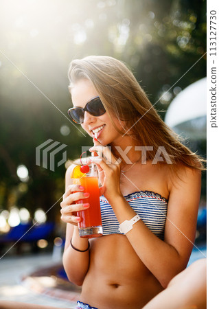 Portrait of young woman with cocktail glass chilling in the tropical sun near swimming pool on a deck chair with palm trees behind. Vacation concept Portrait of young woman with cocktail glass chilling in the tropical sun near swimming pool on a deck chair with palm trees behind. Vacation concept 113127730