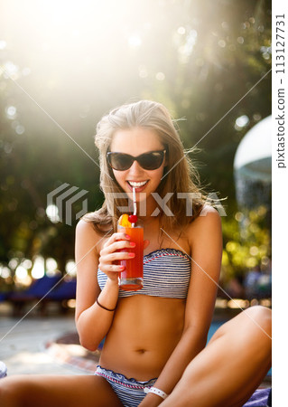 Portrait of young woman with cocktail glass chilling in the tropical sun near swimming pool on a deck chair with palm trees behind. Vacation concept 113127731