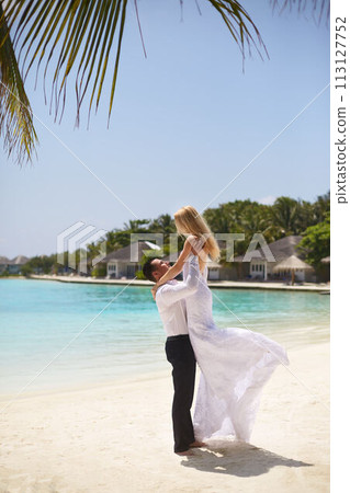 Happy groom holds bride on his hands under wedding ceremony arch on tropical island beach on Maldives. Turquoise ocean lagoon, white sand and bungalows of luxury spa resort on background. Honeymoon. 113127752