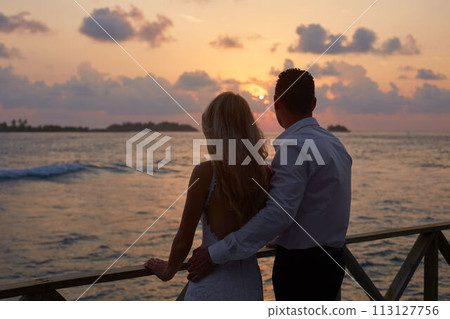 Back view of newly married hug each other and watch sunset skyline on tropical island beach on Maldives. Turquoise ocean lagoon on background. Honeymoon concept. 113127756