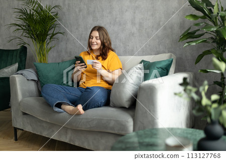 Cheerful young woman in casual clothes sit on couch spending time in living room at home. Rest relax good mood leisure lifestyle concept. 30s female Using phone, hold credit bank card, full body 113127768