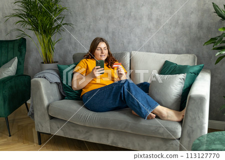 Cheerful young woman in casual clothes sit on couch spending time in living room at home. Rest relax good mood leisure lifestyle concept. 30s female Using phone, hold credit bank card, full body Cheerful young woman in casual clothes sit on couch spending time in living room at home. Rest relax good mood leisure lifestyle concept. 30s female Using phone, hold credit bank card, full body 113127770