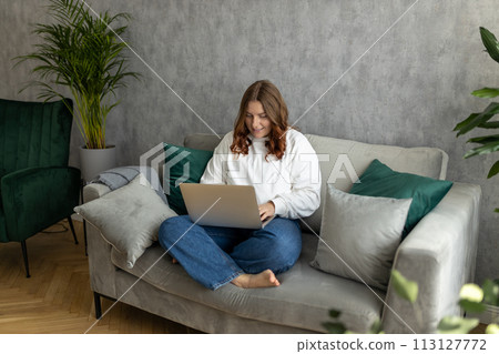 Nice beautiful 30s woman with red hair working at the notebook sitting on the sofa at home. Concepts of education, work and comfort. Full body 113127772