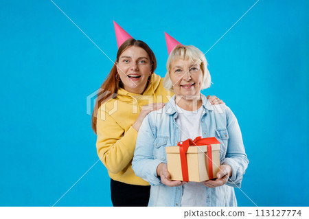 Young smiling happy daughter and mother together gifting birthday present with red ribbon isolated on blue color background studio. Human emotions, facial expression and holiday concept. 113127774
