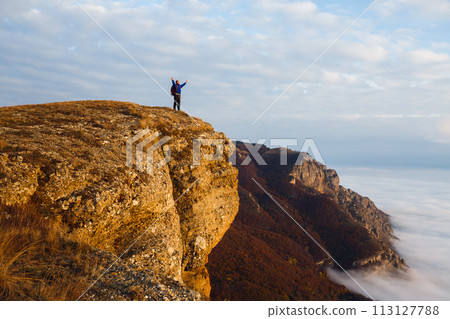 Hiker man cheering with arms spread and raised to the sky after hiking mountain top summit. Climber with backpack on the peak above the clouds. Winner, success, freedom concept. Hiker man cheering with arms spread and raised to the sky after hiking mountain top summit. Climber with backpack on the peak above the clouds. Winner, success, freedom concept. 113127788