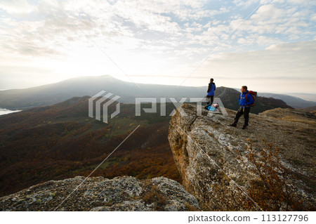 Two hikers with backpacks stand on the top of the mountain and looking at beautiful yellow autumn landscape sunset over clouds. Climbers relax on summit 113127796