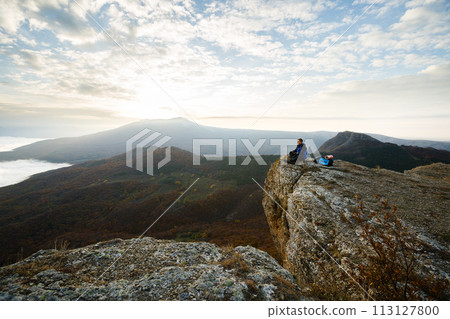 Smiling man hiker with backpack siting and relaxing on the top of the mountain and looking at beautiful yellow autumn landscape sunset over clouds Smiling man hiker with backpack siting and relaxing on the top of the mountain and looking at beautiful yellow autumn landscape sunset over clouds 113127800