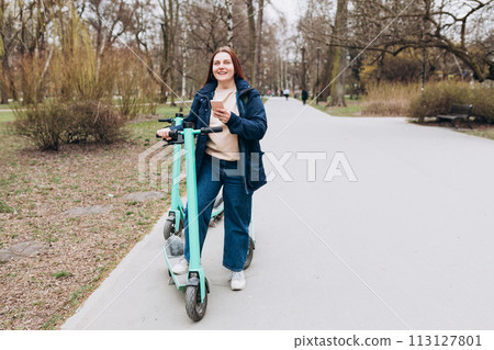 Smiling young woman standing near electric push scooter using smart phone. Ecological transport. Active lifestyle. Urban lifestyle concept. Full body Smiling young woman standing near electric push scooter using smart phone. Ecological transport. Active lifestyle. Urban lifestyle concept. Full body 113127801