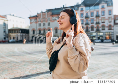 Happy young Caucasian woman in good mood listening to music with headphones and standing on the street in the city. Music lover enjoying music. Urban lifestyle concept. Happy young Caucasian woman in good mood listening to music with headphones and standing on the street in the city. Music lover enjoying music. Urban lifestyle concept. 113127805