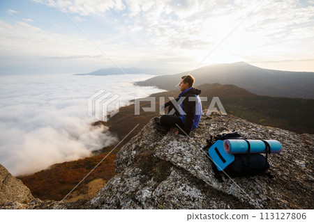 Young tourist hiker with backpack siting and relaxing on the top of the mountain and looking at beautiful yellow autumn landscape sunset over clouds 113127806