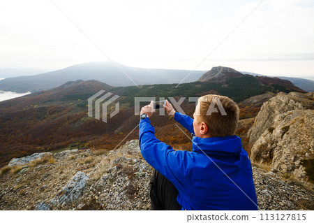 Hiker sitting and taking photo of beautiful mountain landscape with mobile phone. Climber man photographing panorama with smartphone on summit. 113127815