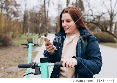 Smiling young woman standing near electric push scooter using smart phone. Ecological transport. Active lifestyle. Urban lifestyle concept. 113127821