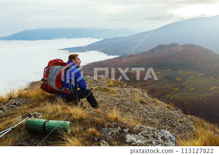 Young tourist hiker with backpack siting and relaxing on the top of the mountain and looking at beautiful yellow autumn landscape sunset over clouds Young tourist hiker with backpack siting and relaxing on the top of the mountain and looking at beautiful yellow autumn landscape sunset over clouds 113127822