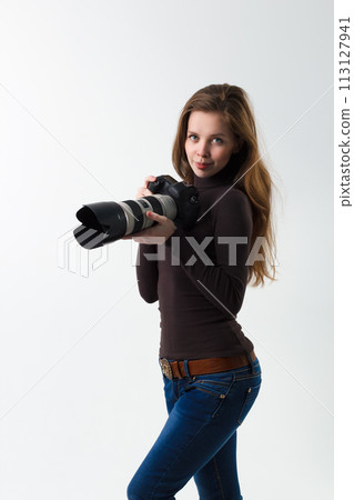 The beautiful photographer girl with professional dslr camera posing on a white background in studio. Photo learning, studying, training concept The beautiful photographer girl with professional dslr camera posing on a white background in studio. Photo learning, studying, training concept 113127941