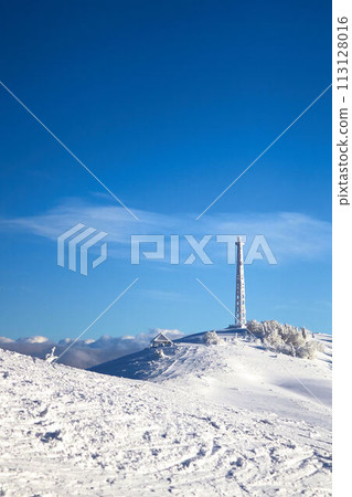 Icy cellular base station antenna covered with snow. Cell site tower on moutain hill. Telephone network transceivers and communication equipment on frosty weather day after snowstorm. Blue sky. 113128016