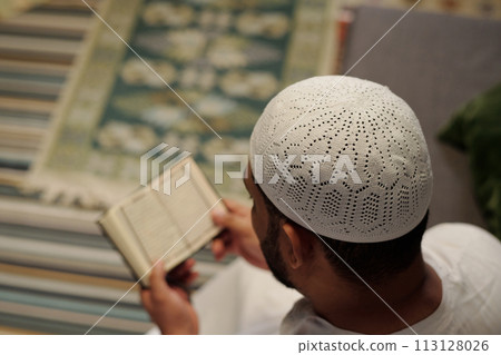 High angle view selective focus shot of unrecognizable Muslim man wearing white taqiyah reading Quran, copy space 113128026