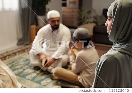 Selective focus high angle shot of young Muslim woman wearing hijab sitting in living room at family gathering Selective focus high angle shot of young Muslim woman wearing hijab sitting in living room at family gathering 113128040