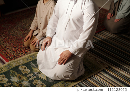 High angle view of unrecognizable Muslim family of three sitting on floor indoors praying salah, copy space High angle view of unrecognizable Muslim family of three sitting on floor indoors praying salah, copy space 113128053