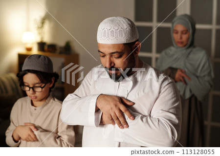 Selective focus shot of bearded Muslim man, his wife in hijab and son standing in living room praying salah Selective focus shot of bearded Muslim man, his wife in hijab and son standing in living room praying salah 113128055