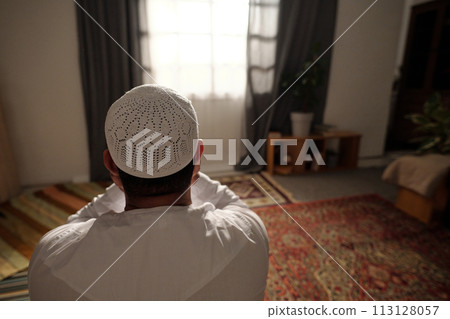 Rear view shot of unrecognizable male Muslim wearing white thobe with taqiyah praying salah in living room at home, copy space Rear view shot of unrecognizable male Muslim wearing white thobe with taqiyah praying salah in living room at home, copy space 113128057