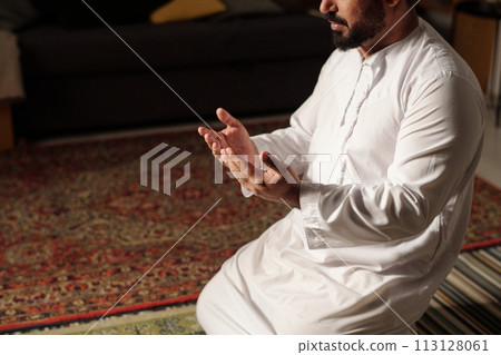 High angle view shot of bearded Muslim man in white clothes sitting on rug praying salah, copy space 113128061