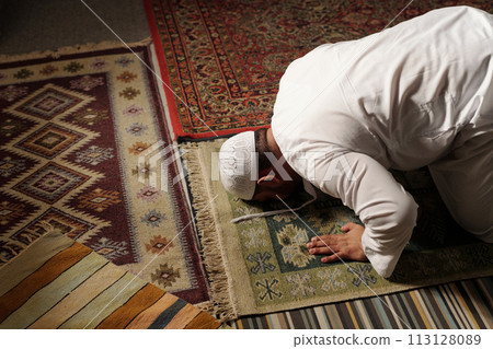 High angle view of mature Muslim man wearing white thobe and tuqiyah doing sajdah on prayer rug during namaz, copy space 113128089