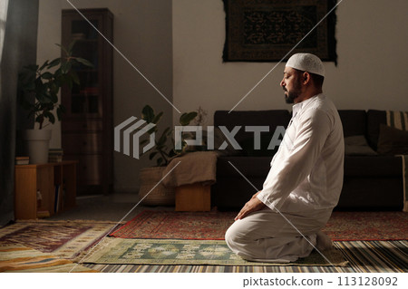 Side view shot of mature Muslim man wearing white clothes sitting on prayer rug in living room doing namaz 113128092