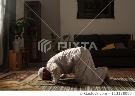 Long shot of mature bearded Muslim man wearing white thobe doing sajdah on prayer rug in living room at home, copy space 113128093
