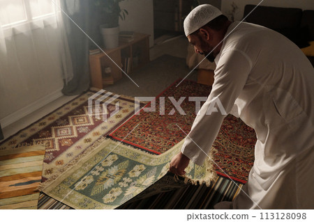 High angle view of Muslim man laying green prayer rug on floor in living room before doing namaz, copy space High angle view of Muslim man laying green prayer rug on floor in living room before doing namaz, copy space 113128098