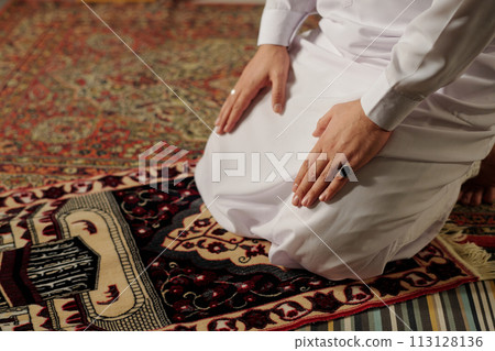 High angle view of unrecognizable young Muslim man wearing white thobe doing namaz on prayer rug, copy space 113128136