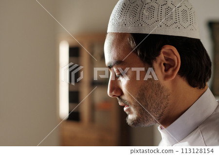 Side view closeup of young Muslim man wearing white taqiyah on head standing with eyes closed praying salah, copy space 113128154