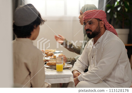 Selective focus shot of Muslim man wearing kandora and keffiyeh sitting festive dining table chatting with his nephew during family dinner 113128176