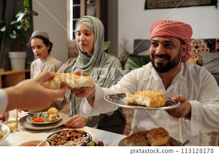 Selective focus shot of cheerful Muslim man sitting at table sharing flatbread during Uraza Bayram celebration dinner 113128178