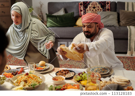 Muslim man wearing keffiyeh tearing off piece of flatbread during festive dinner with family on Eid Al-Fitr 113128183