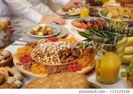 Selective focus shot of sweet dried fruit and nuts on wooden platter on festive dining table, unrecognizable Muslim people celebrating Eid Al-Fitr 113128186