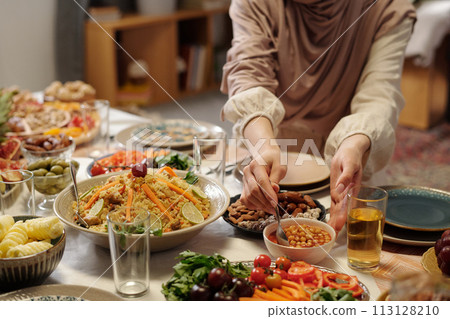 Unrecognizable young Muslim woman wearing hijab setting festive table putting spoon into chickpea dish, copy space 113128210