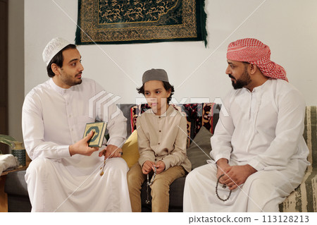 Modern Muslim men and boy sitting on couch in living room discussing suwar from Quran at family gathering on Islamic holiday 113128213