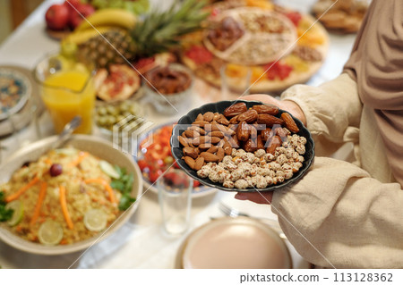 High angle view closeup of hands of unrecognizable Muslim woman holding plate full of almonds, dates and peanuts while setting festive table 113128362