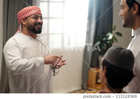 Joyful Muslim man holding prayer beads chatting with his younger brother and nephew at family gathering on Eid Al-Fitr Joyful Muslim man holding prayer beads chatting with his younger brother and nephew at family gathering on Eid Al-Fitr 113128369
