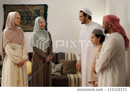 Modern Muslim men, women and kid wearing modest clothes standing in living room having conversation at family gathering on Eid Al-Fitr Modern Muslim men, women and kid wearing modest clothes standing in living room having conversation at family gathering on Eid Al-Fitr 113128372