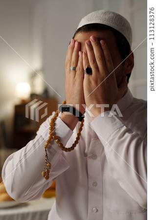 Vertical medium closeup of unrecognizable young Muslim man wearing white clothes with taqiyah covering face with hands while praying salat 113128376
