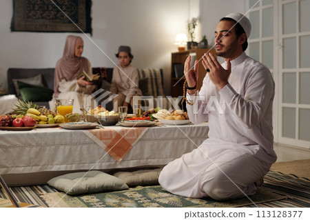 Selective focus shot of young muslim man sitting on floor by festive table praying salah, his wife and son reading Quran in background Selective focus shot of young muslim man sitting on floor by festive table praying salah, his wife and son reading Quran in background 113128377