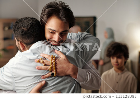 Medium closeup of young Middle Eastern man greeting his friend or relative with hug while gathering with family on Islamic holiday 113128385