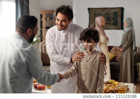 Medium shot of modern Muslim man and his son wearing thobes greeting male relative on Eid al-Fitr, two women talking in background 113128386