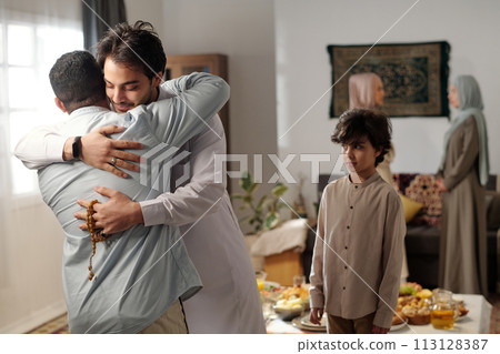 Medium shot of young Muslim man wearing thobe greeting male relative with hug on Eid al-Fitr, his son standing next to him 113128387