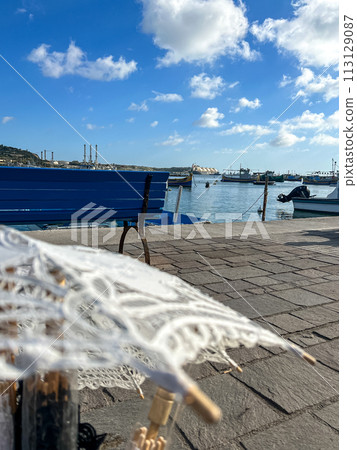 White umbrella on the beach with a view of the sea and boats at Marsaxlokk, Malta 113129087